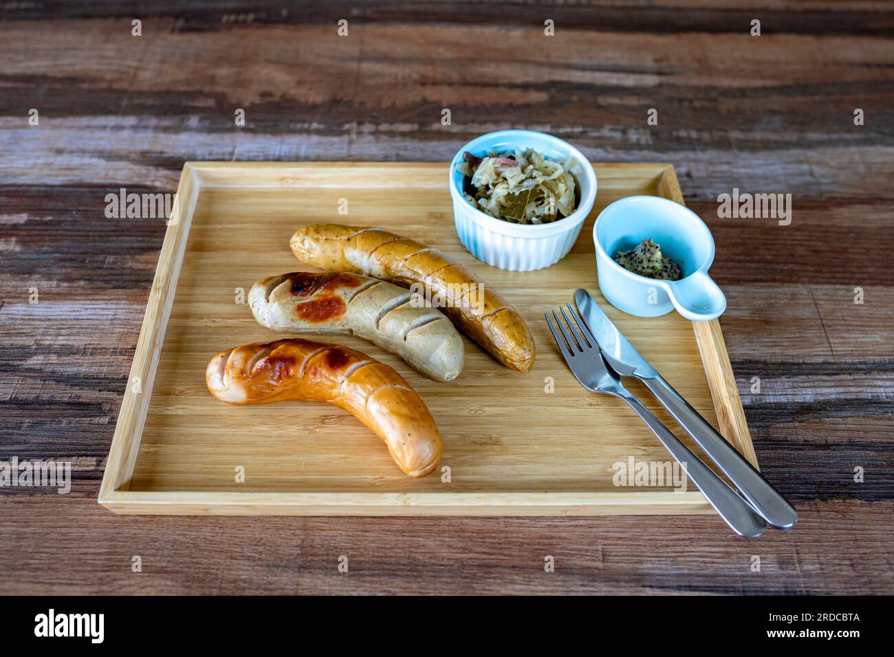 Photos of Grilled German Sausages with sauerkraut and dijon mustard in small bowls with knife and fork Together in a rectangular wooden tray, on a woo Stock Photo