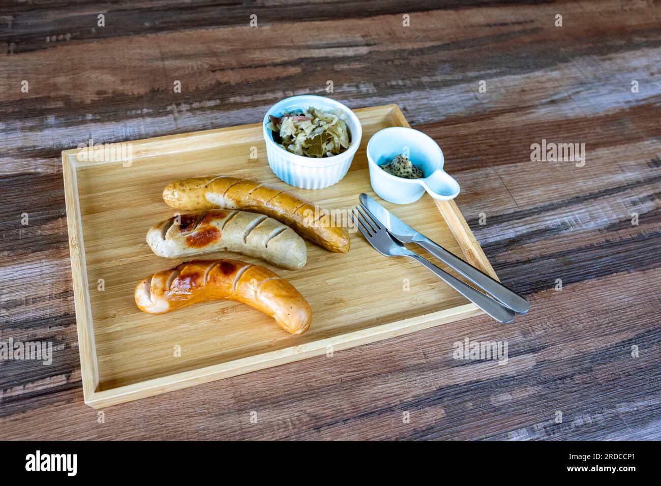Photos of Grilled German Sausages with sauerkraut and dijon mustard in a small bowl with knife and fork Together in a rectangular wooden tray, on a wo Stock Photo