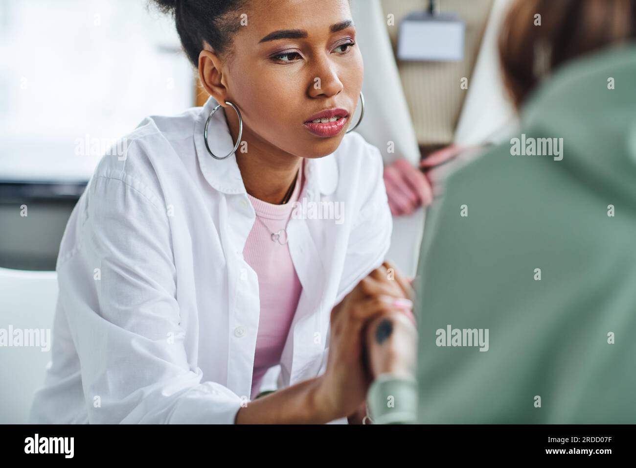 young and serious african american woman holding hands of friend near professional psychologist on blurred background in consulting room, empathy, sol Stock Photo
