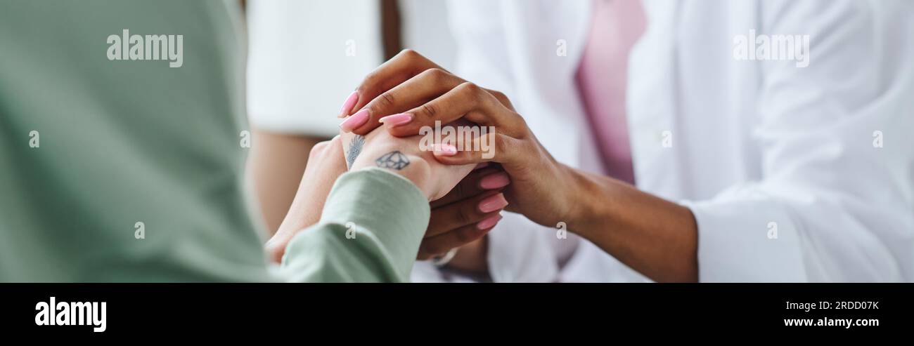 partial view of african american woman holding hands of tattooed female friend while supporting and calming her during psychological therapy, empathy, Stock Photo