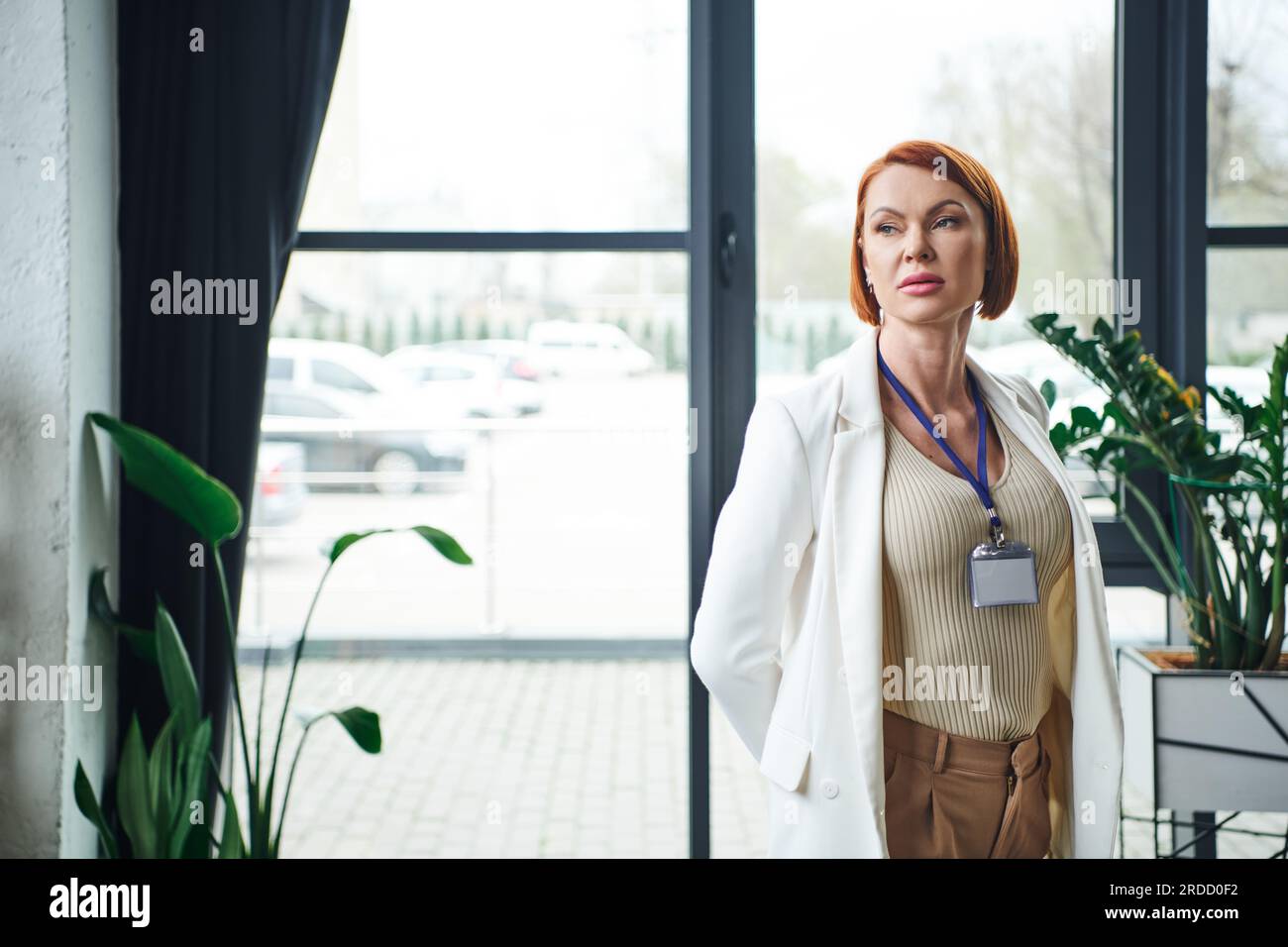 thoughtful psychologist with red hair and name badge standing in white blazer, thinking and looking away in consulting room, professional help and sup Stock Photo
