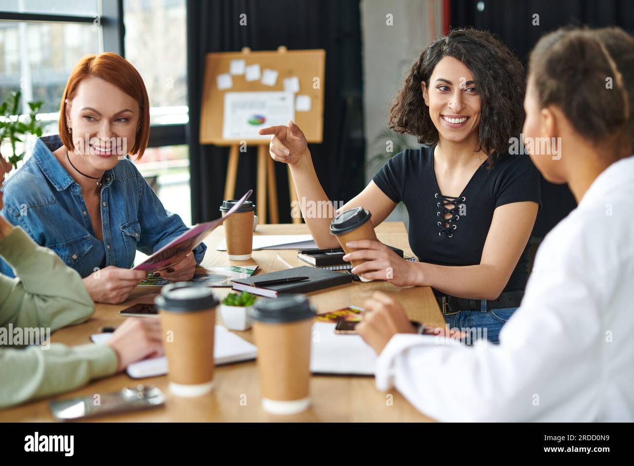 cheerful multiracial woman holding coffee to go and pointing with finger while talking to african american friend near members of interest club, leisu Stock Photo