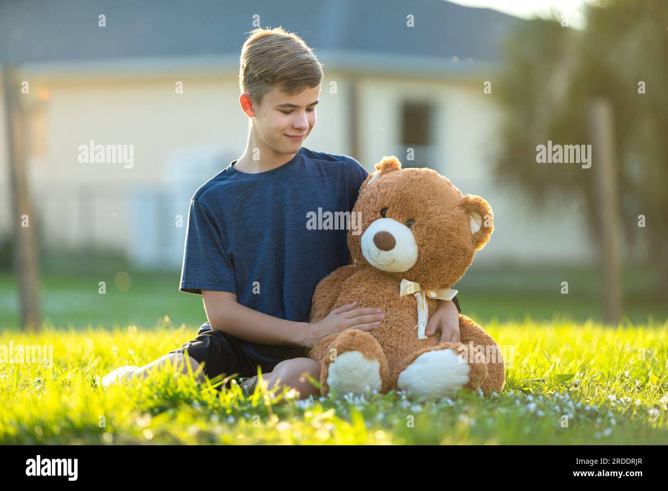 Happy teenager boy hugging his teddy bear friend outdoors on green grass lawn. Friendship concept. Stock Photo