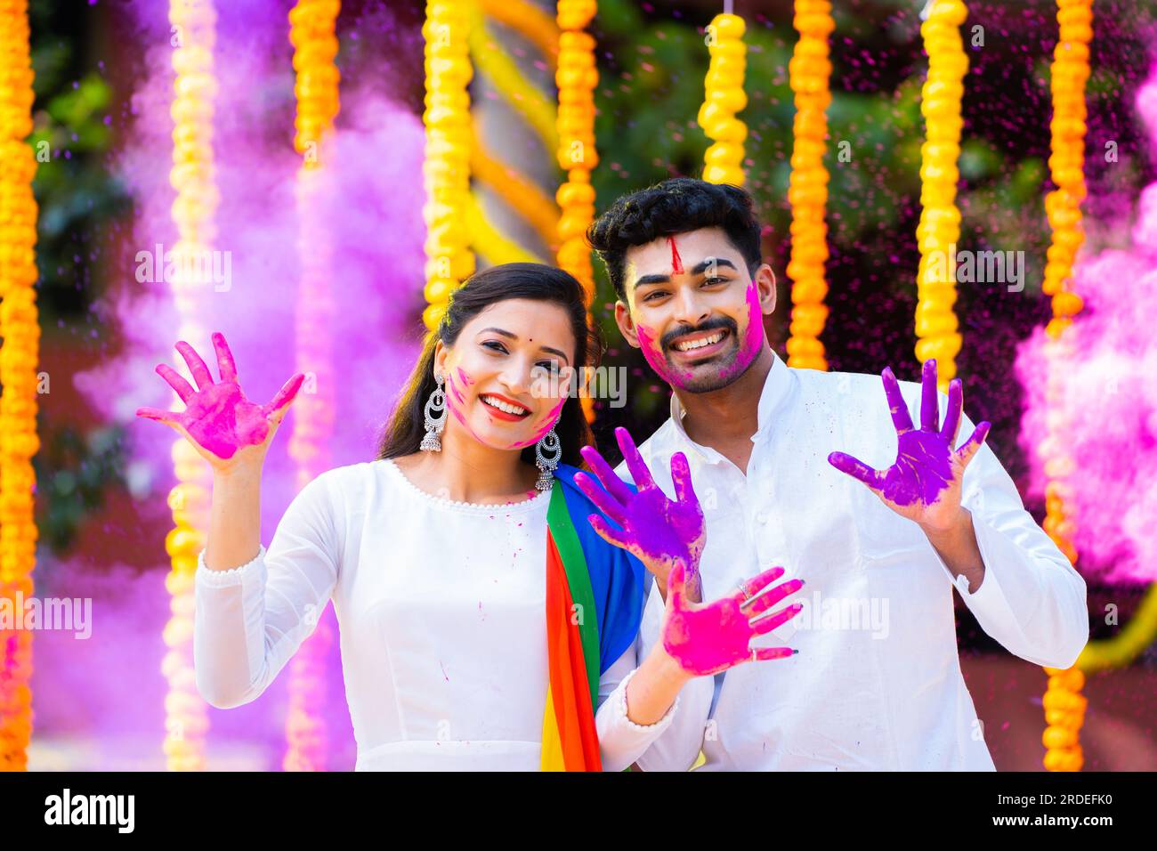 Happy indian young couple showing painted hands by looking at camera while playing holi festival celebration at flower decoration background - concept Stock Photo