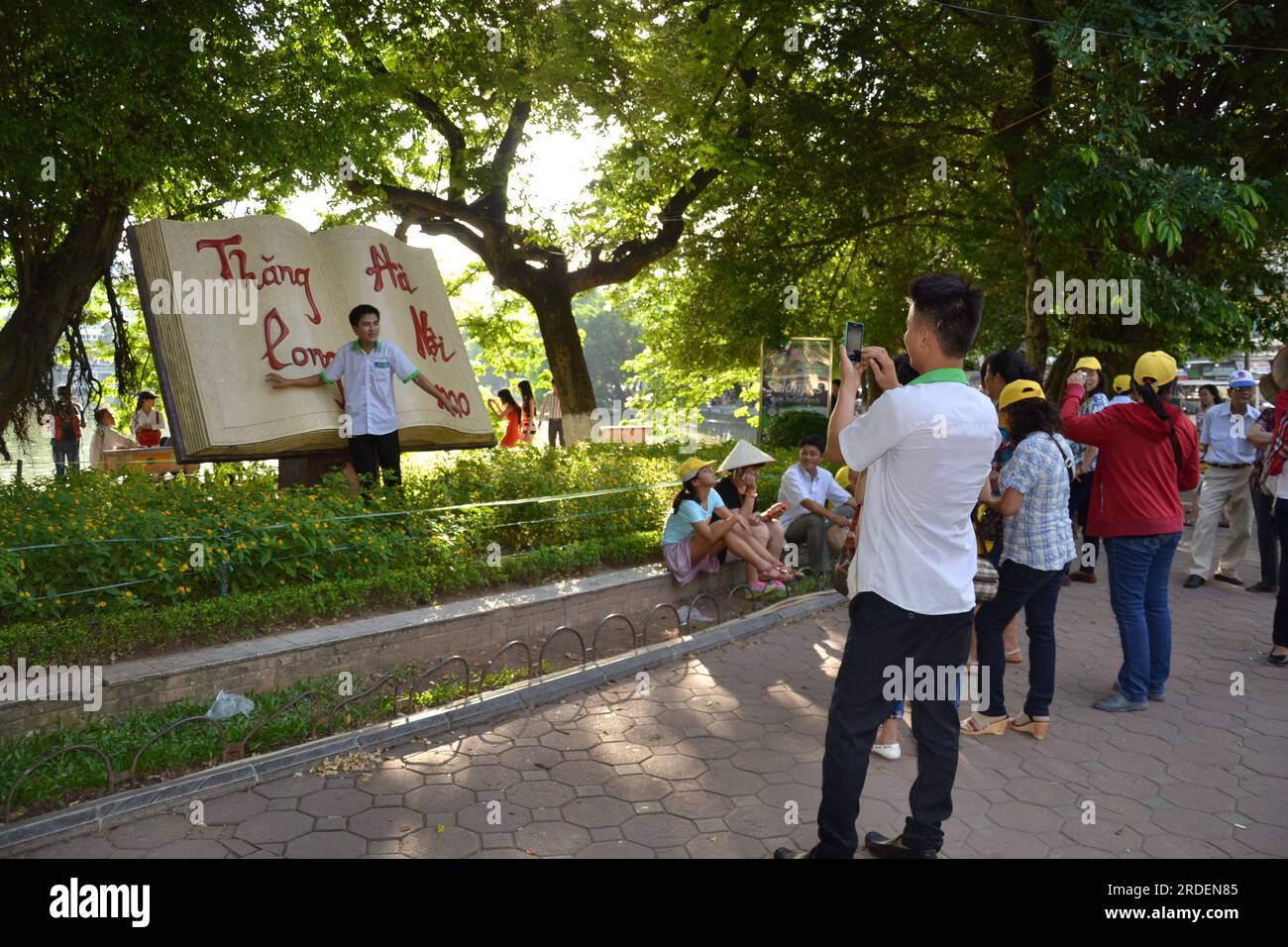 A young man takes a photo of a friend posing in front of the giant Open Book monument in Hoan Kiem Lake park in Hanoi, Vietnam, Asia; giant open book art sculpture celebrates 1000 or one thousand years of culture, founding of Hanoi Stock Photo