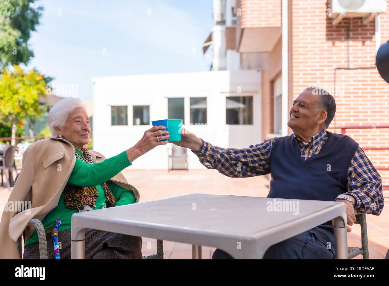 Two elderly people in the garden of a nursing home, toasting with vibrant glasses on a sunny summer day Stock Photo