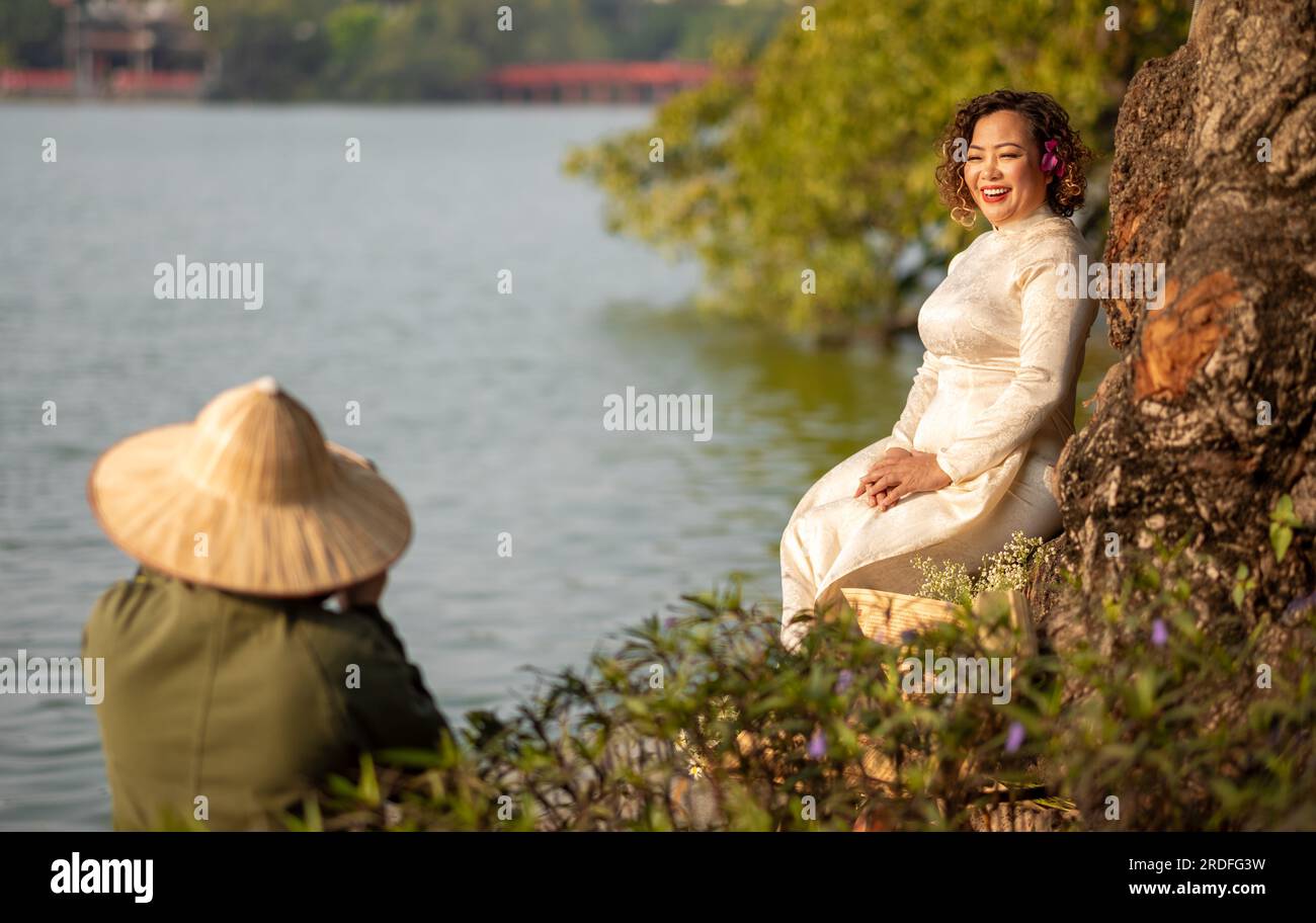 A middle aged Vietnamese woman wearing a traditional 'Ao Dai' poses for a photograph next to Hoan Kiem Lake in central Hanoi, Vietnam. Stock Photo