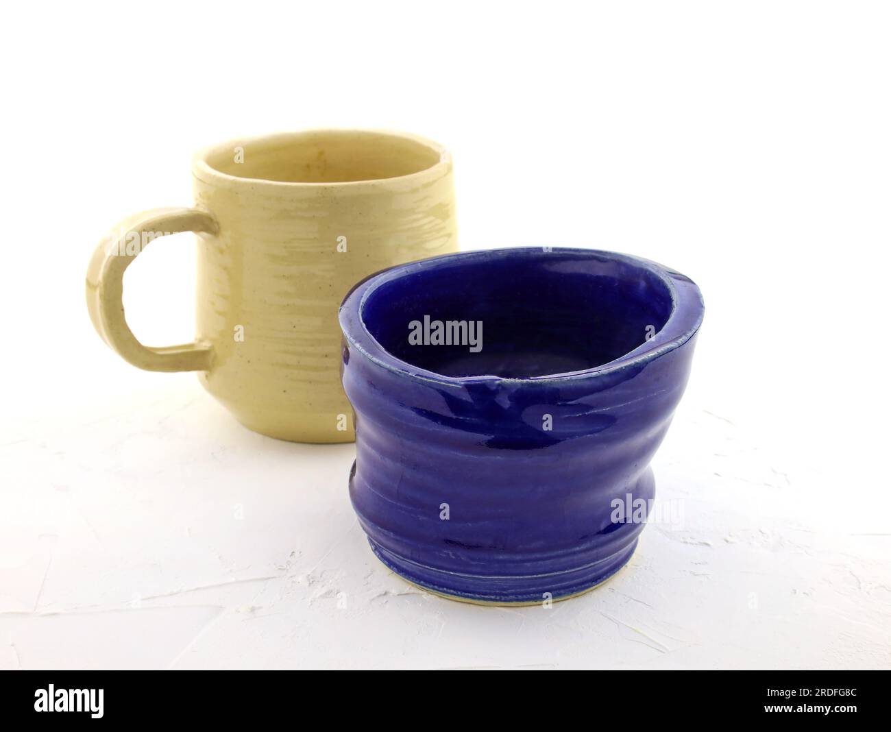 Color ceramic dishes on a white background. Cup made of blue clay covered with glaze and beige cup with handle. Empty cups in stack. ceramic products Stock Photo