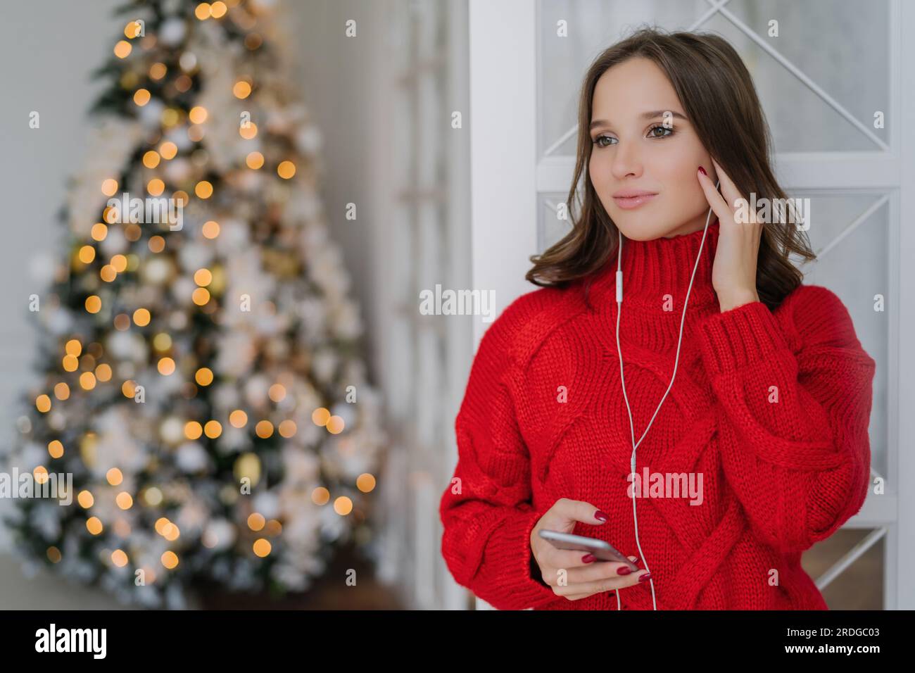Tech-savvy woman enjoys holidays, uses phone and earphones near Christmas tree. Copy space available. Stock Photo