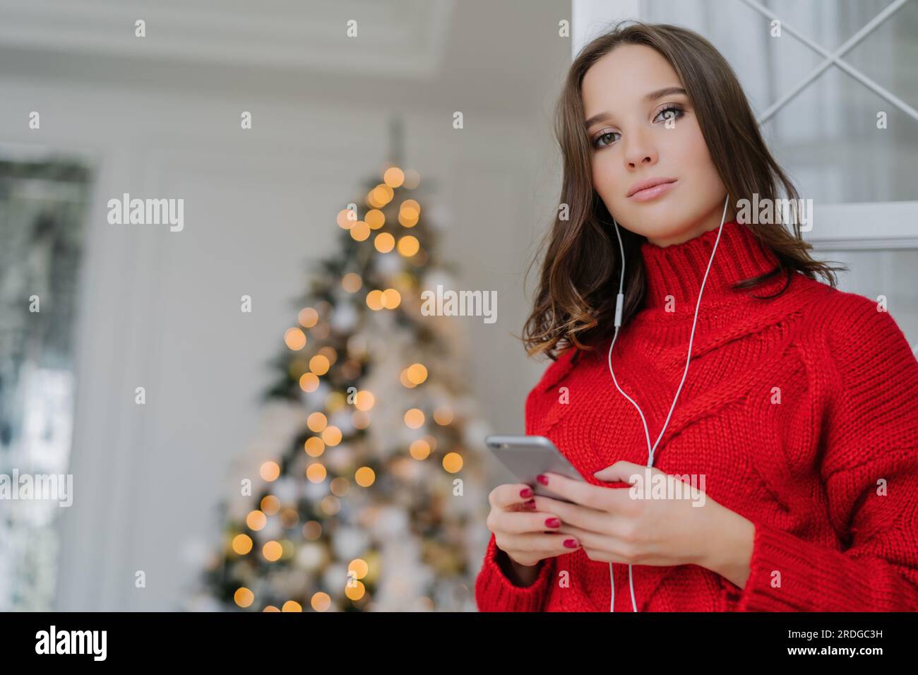 European girl with makeup, healthy skin, in oversized sweater, listens to audio book by Christmas tree. Stock Photo