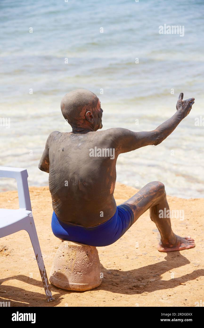 Man covered in mineral mud rub gesticulating whilst sat on the shore, The Dead Sea, Jordan Stock Photo