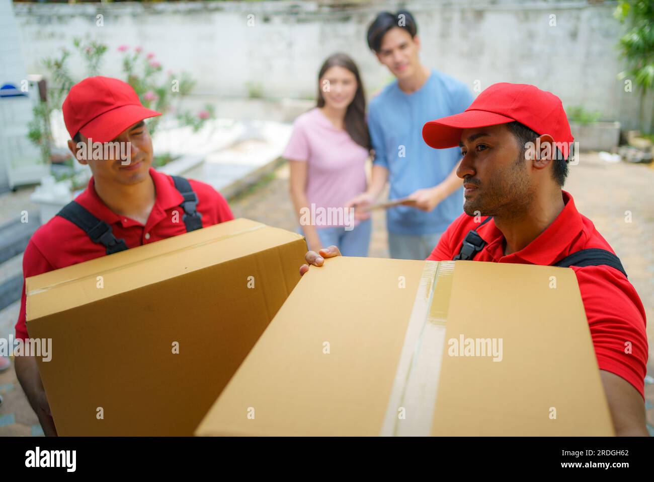 An Asian couple stands together, observing with anticipation as workers move into their new home. Excitement fills the air as they eagerly anticipate Stock Photo