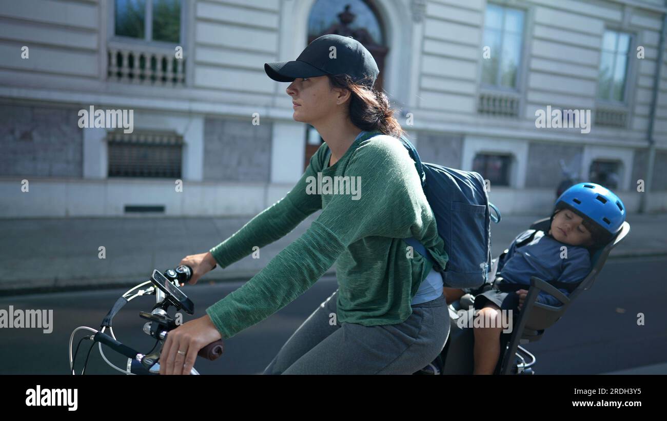 Mother riding bicycle in street with child in back seat asleep Stock