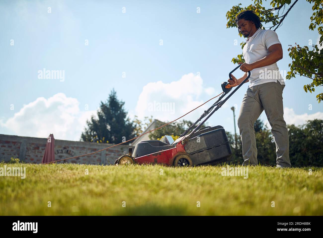 Handsome young african american man mowing grass outdoors, making the backyard beutiful and neat. Lifestyle, activity, home concept. Stock Photo