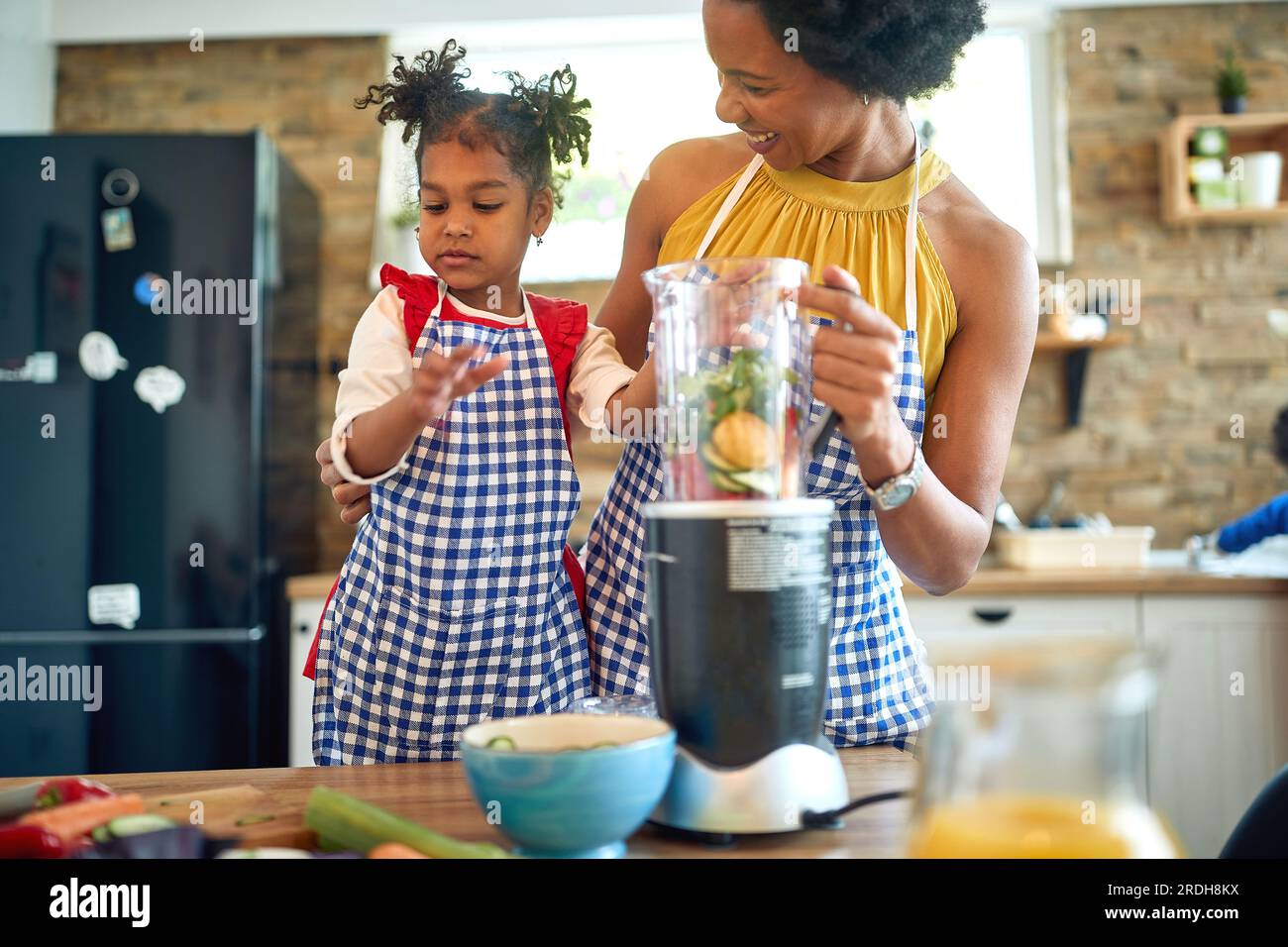 Mother and her young daughter coming together to prepare a healthy meal. Immersed in the process, they are seen adding fresh vegetables to a blender t Stock Photo