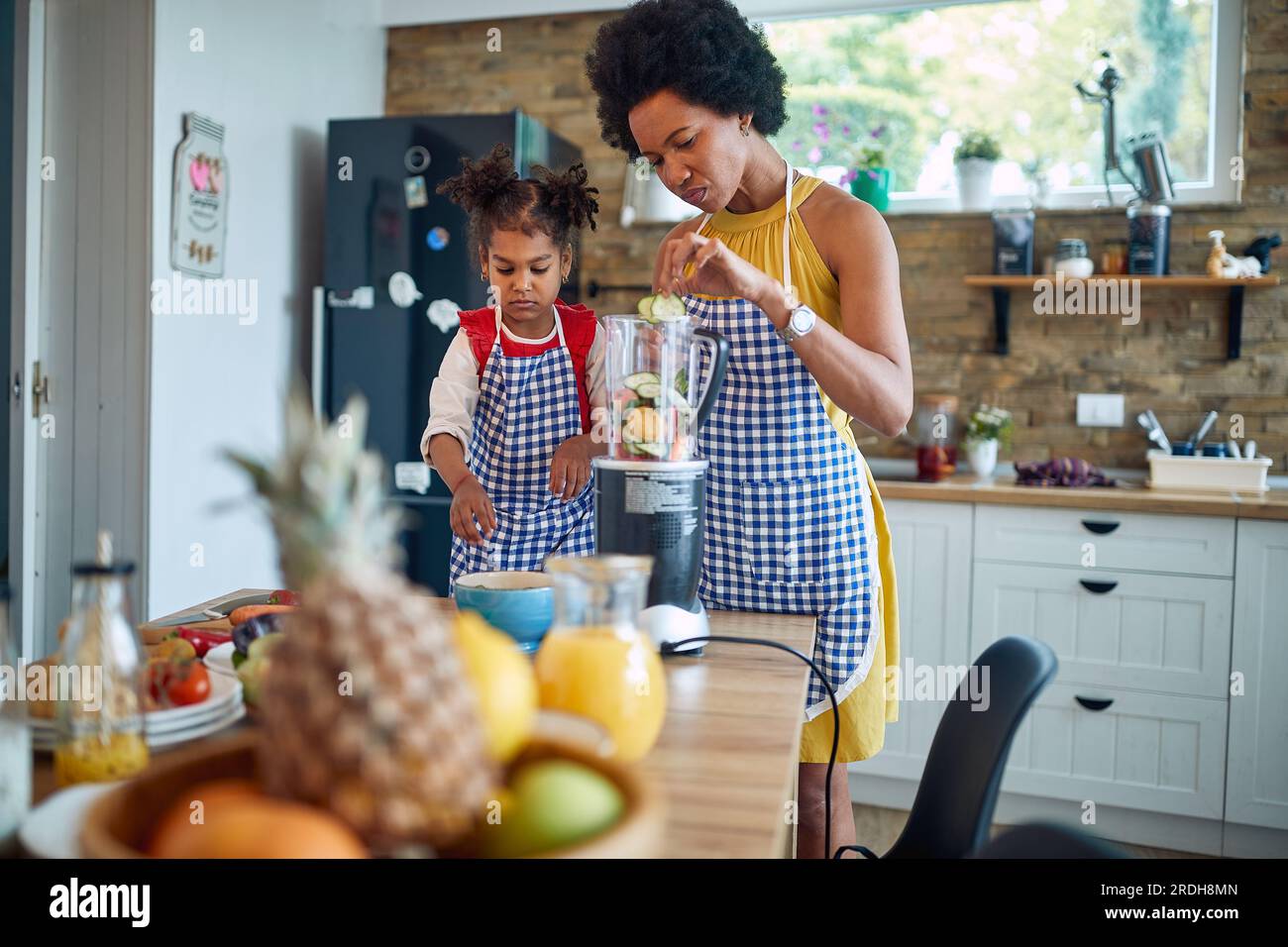 Mother and her young daughter coming together to prepare a healthy meal. Immersed in the process, they are seen adding fresh vegetables to a blender t Stock Photo