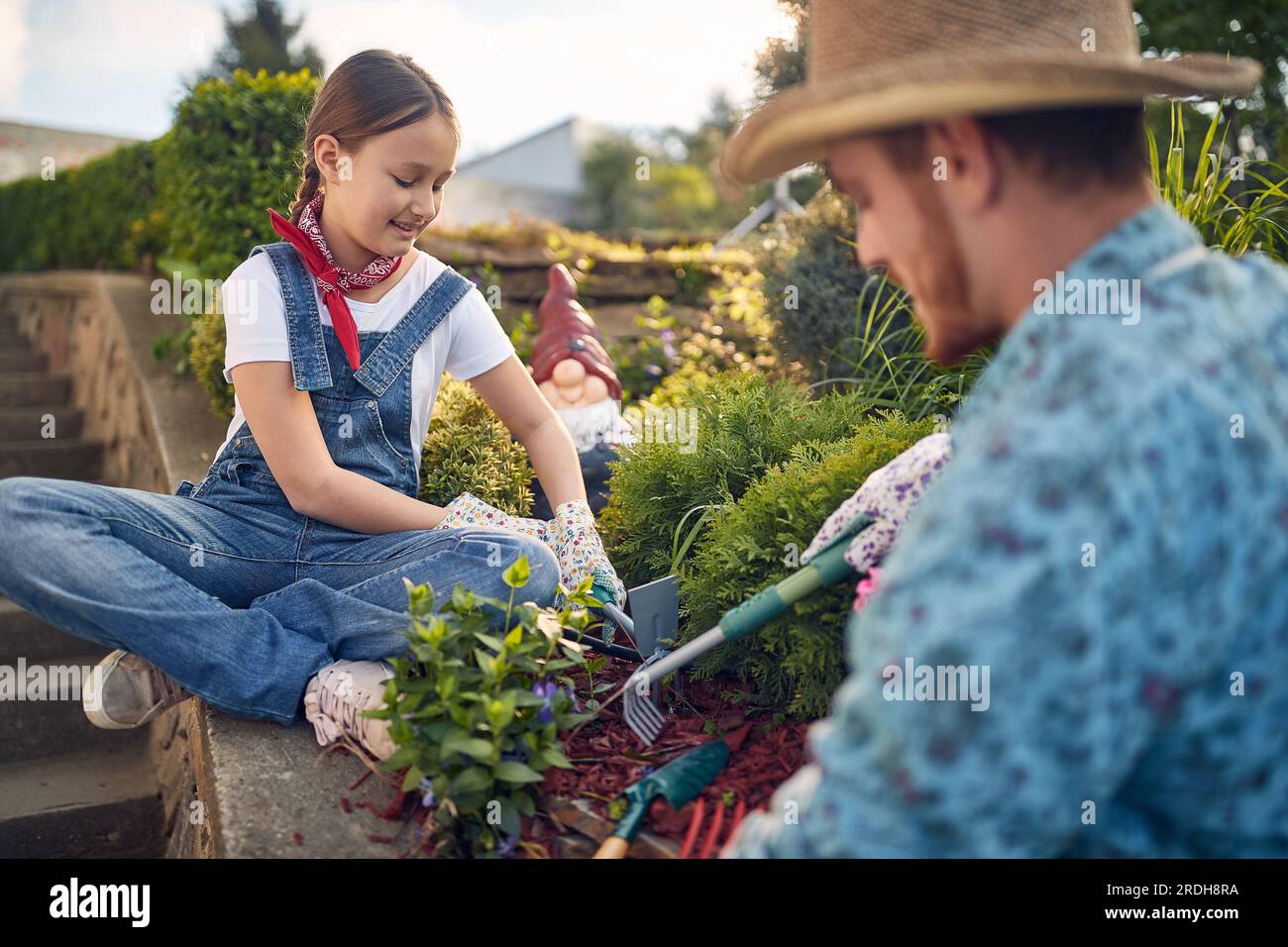 Beautiful young girl using garden rake sitting by the garden outdoors planting flowers with her father on a sunny summer day. Home, lifestyle, family Stock Photo