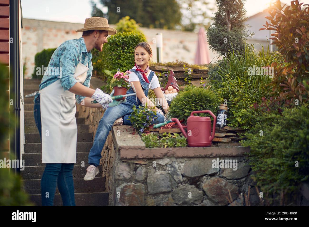 Happy family working outdoors. Young father and girl planting flowers in the flower garden by the house on a sunny summer day, feeling cheerful. Home, Stock Photo