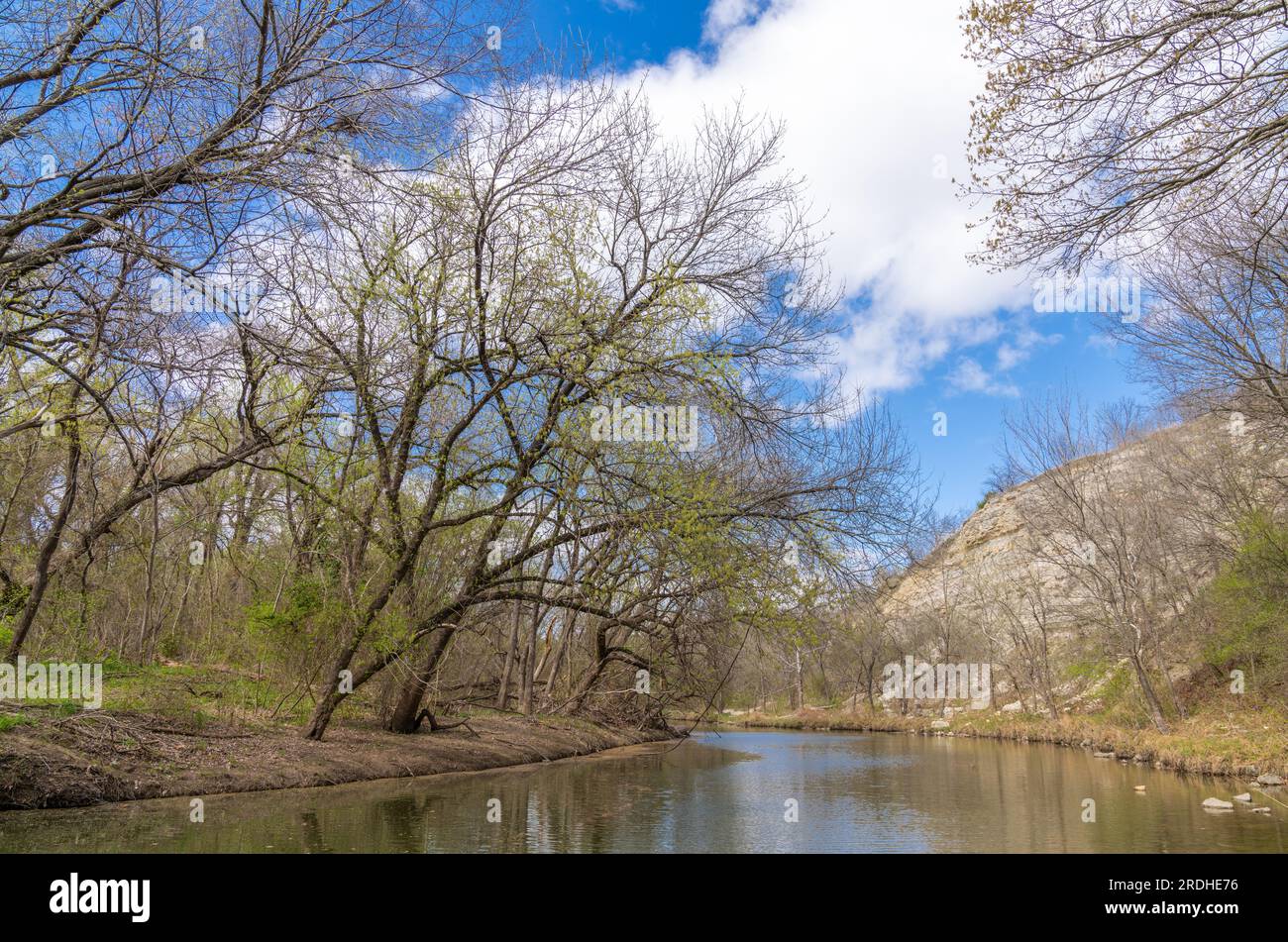 Partly cloudy early Spring day in the woods with a quiet and tranquil flowing stream flanked on one side with a towering, rocky, limestone hillside Stock Photo