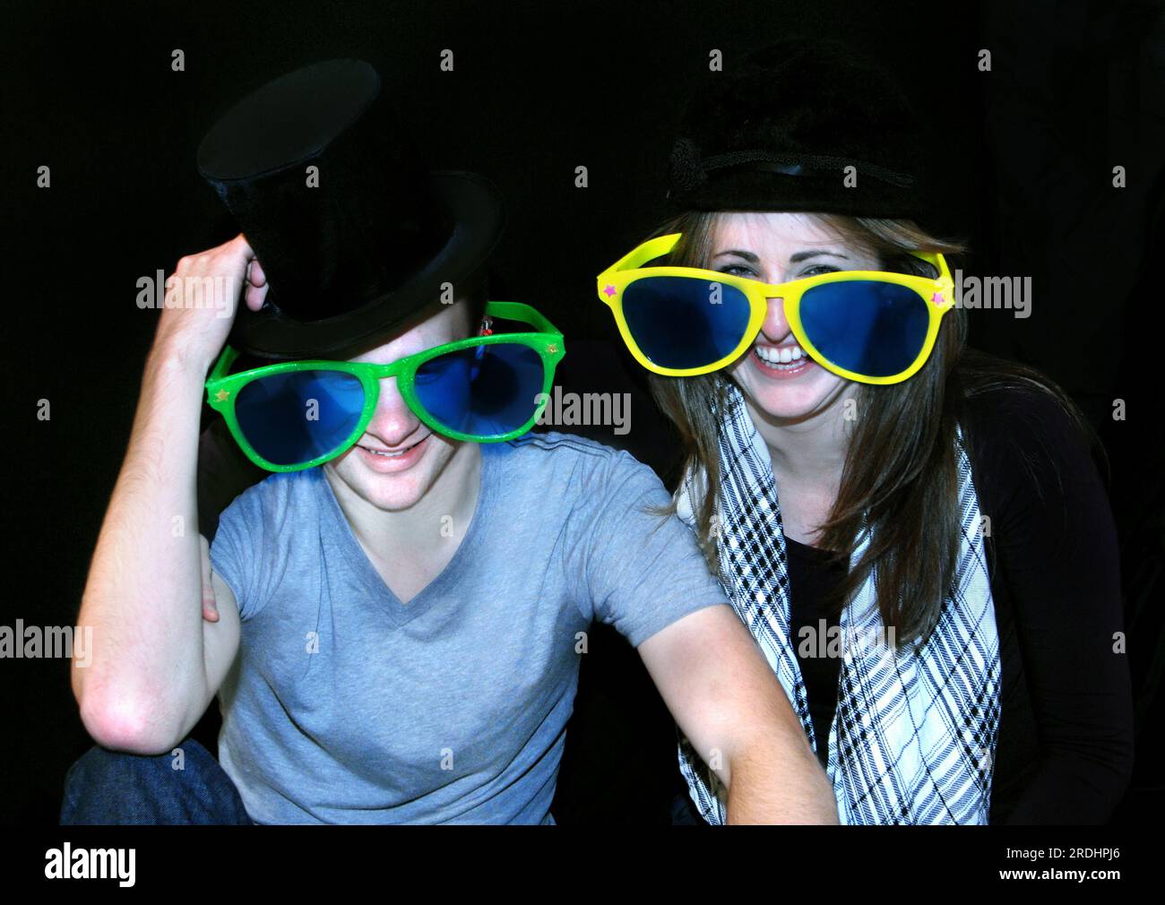 Young couple clown around with hats and oversized sunglasses.  They are both smiling and the man is tipping his top hat. Stock Photo
