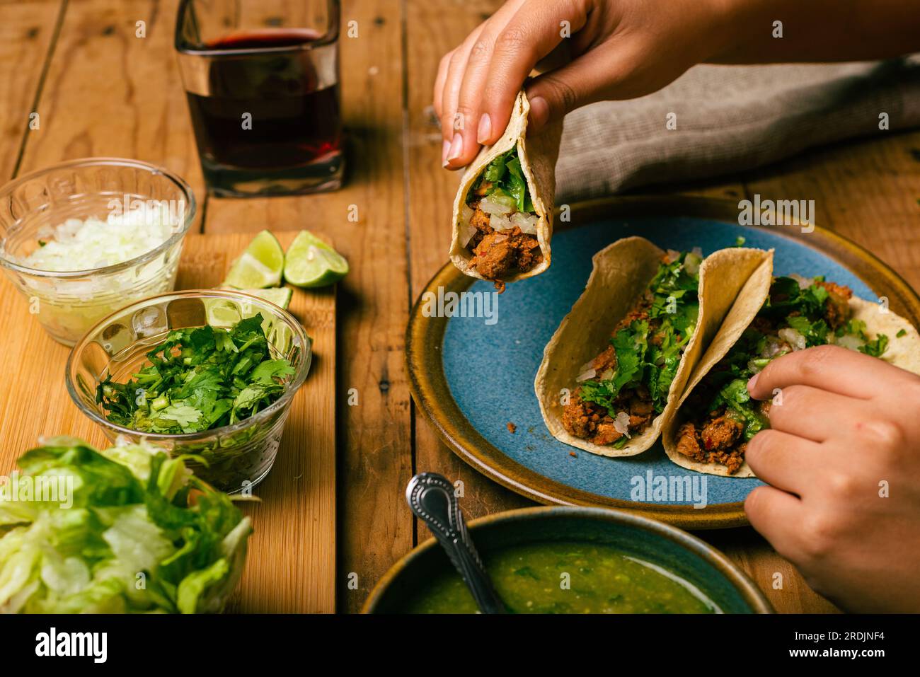Woman's hand holding a taco of marinated meat. Plate with tacos, sauce and vegetables on wooden table. Top view. Stock Photo