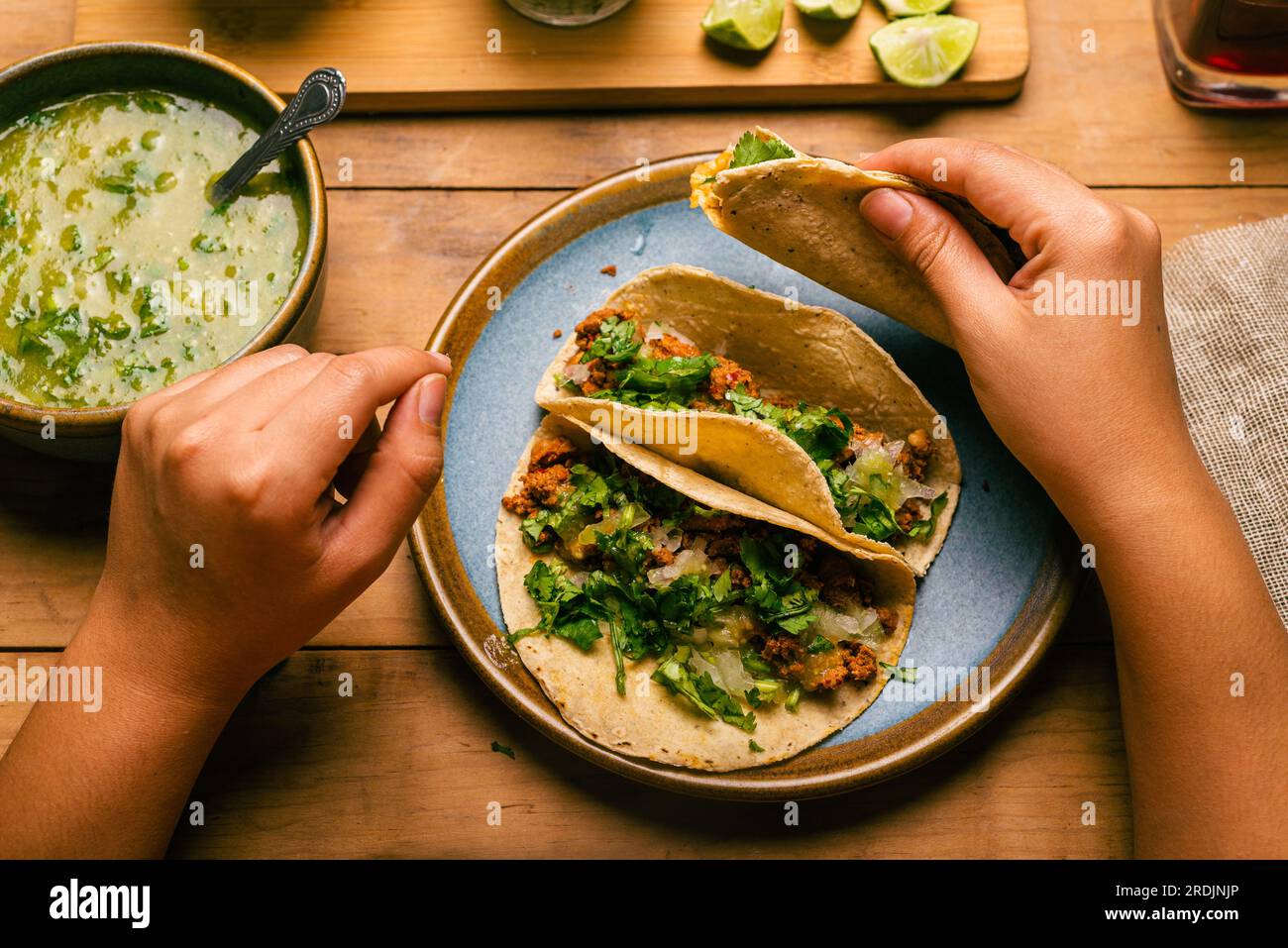 Woman's hand holding a taco of marinated meat. Plate with tacos, sauce and vegetables on wooden table. Top view. Stock Photo