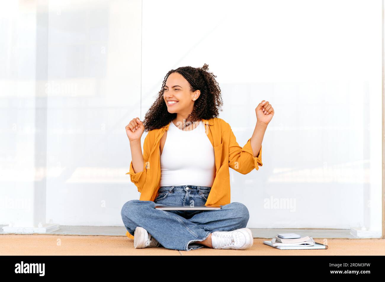 Finishing work or study. Joyful curly haired latino or brazilian female student, sits on the floor near the university, finished studying online, rejoicing, gesturing with hands, dance, smiling Stock Photo