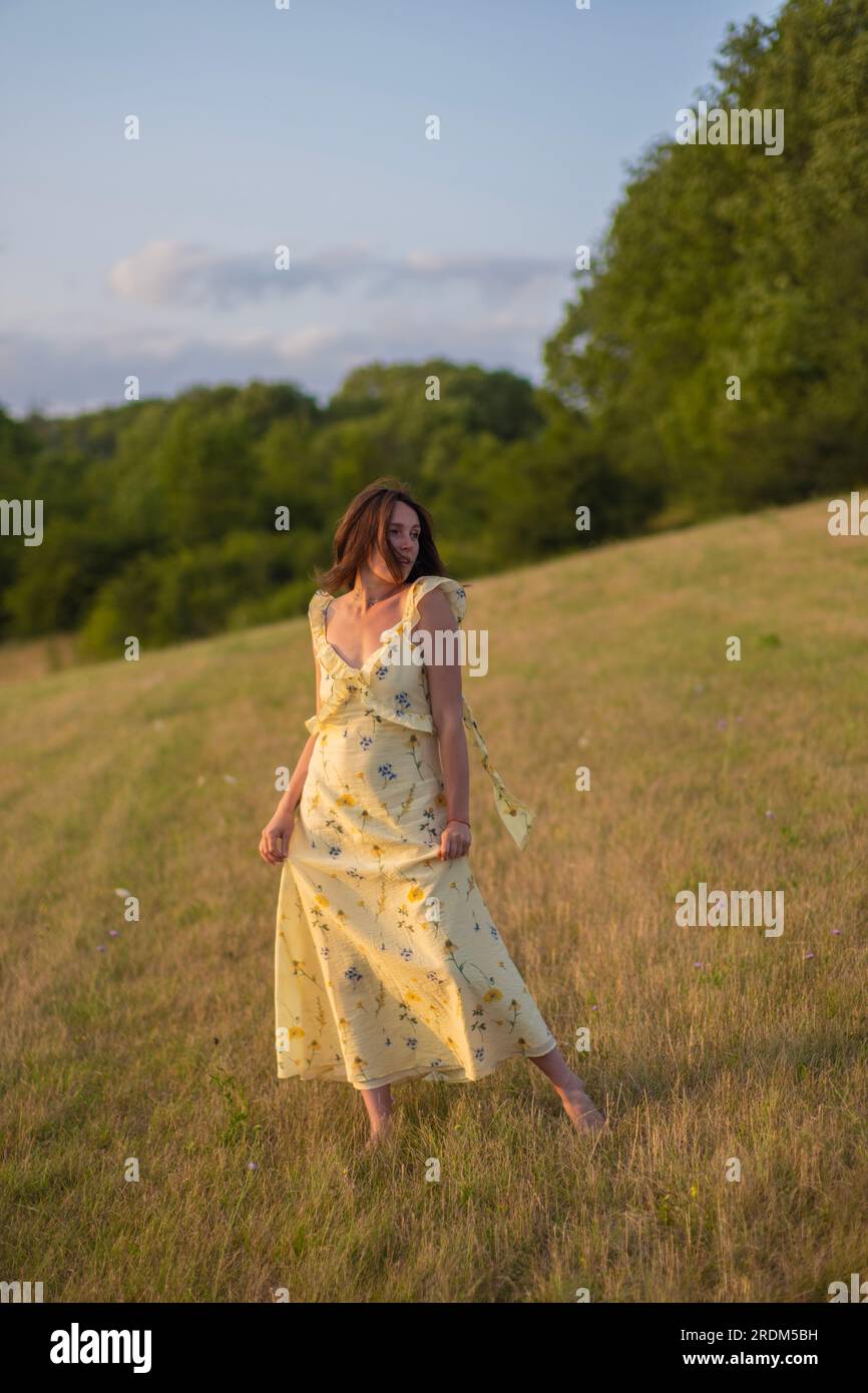 A young beautiful lady walks barefoot in a field at sunset. A girl in a beautiful yellow dress poses at sunset in a field. Stock Photo