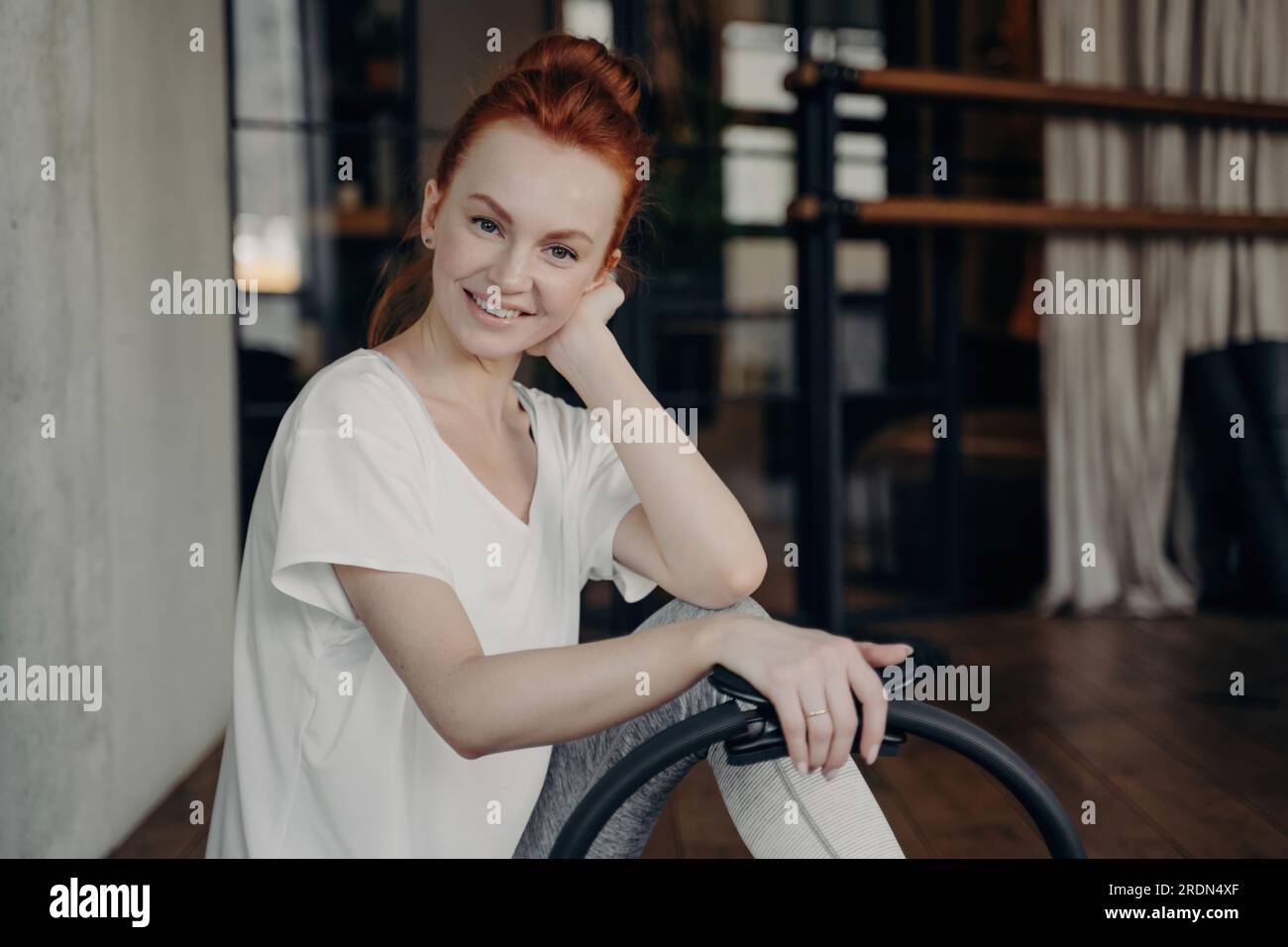 Gorgeous ginger female fitness instructor in white t-shirt smiles at camera during Pilates ring workout in gym. Promoting a healthy, active lifestyle. Stock Photo