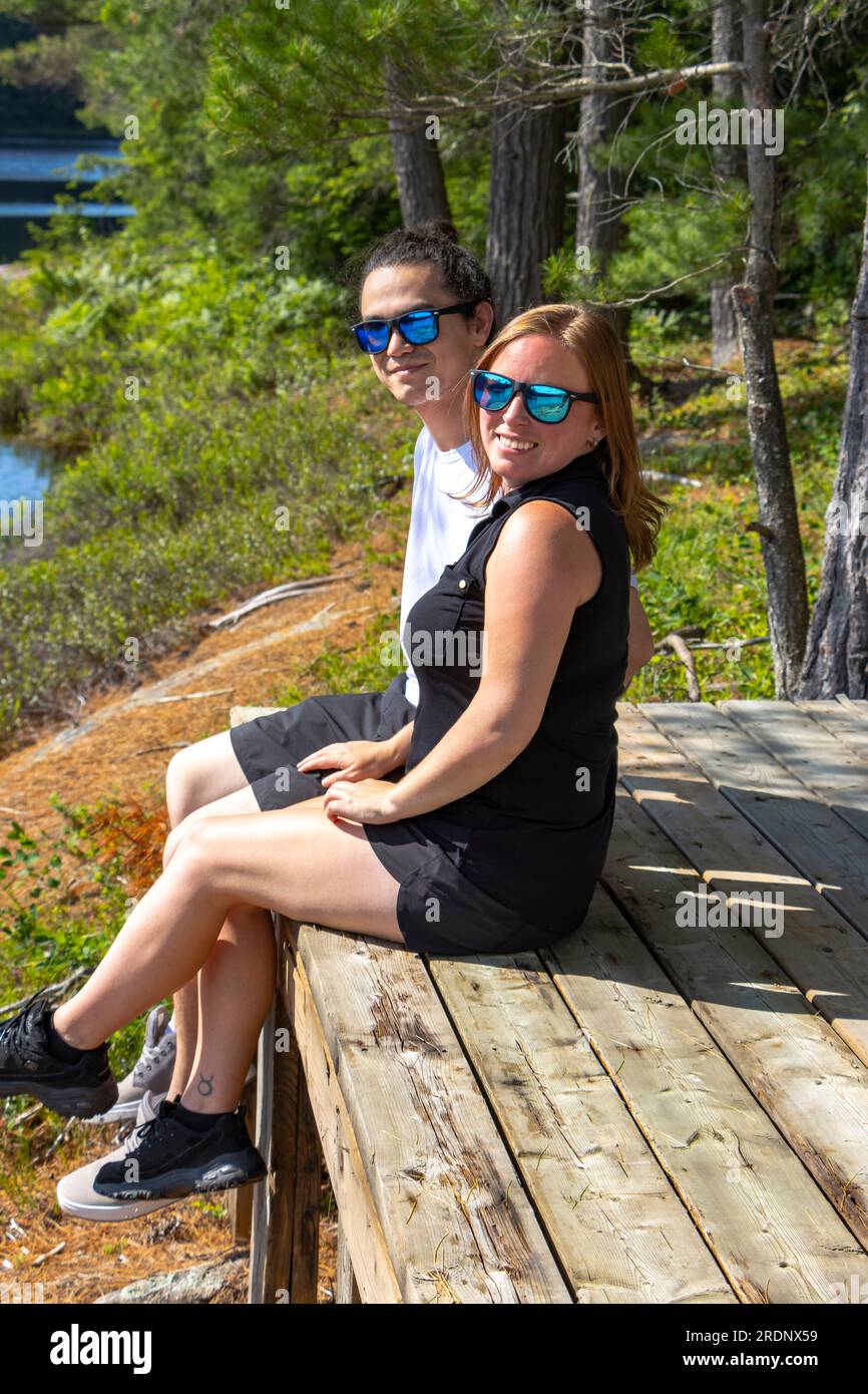 mixed race couple sitting on dock smiling at camera in Muskoka forest Stock Photo