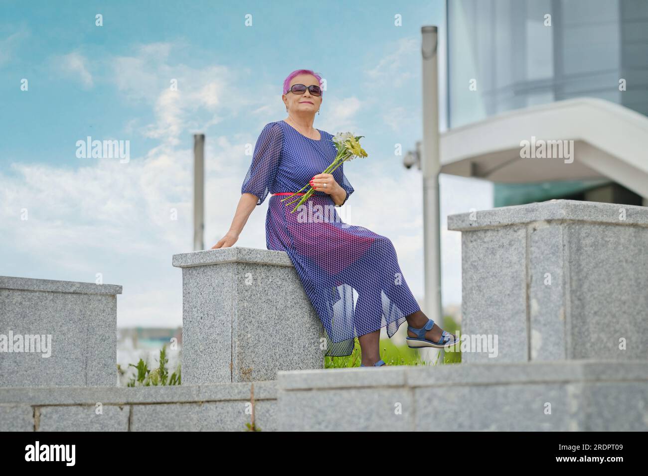 Outdoor portrait of smiling mature woman with short pink hair with spring flowers in hands Stock Photo