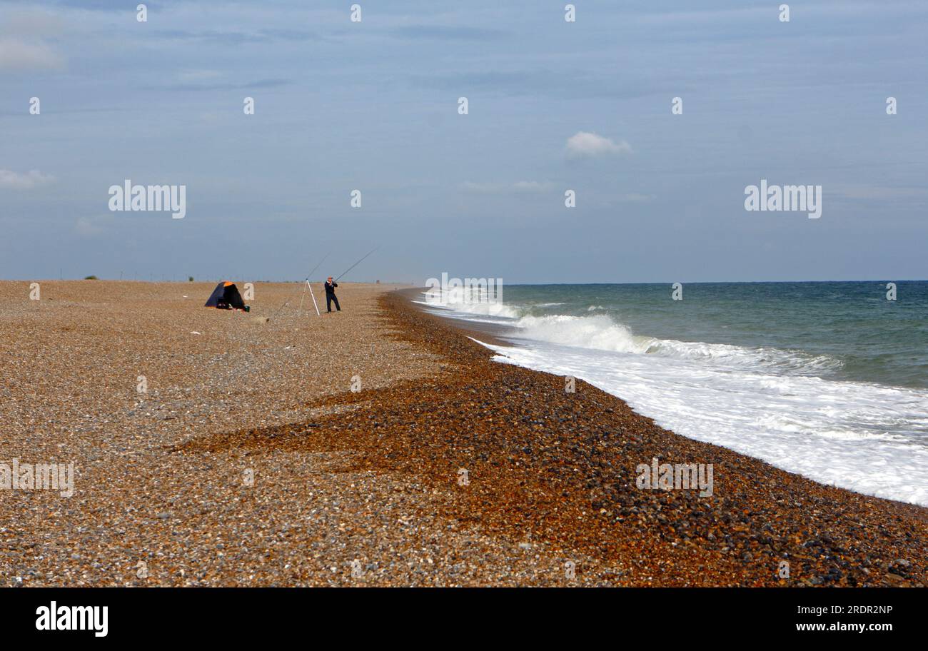 A sea angler on the shingle beach with moderate sea on a windy day in summer in North Norfolk at Cley Next the Sea, Norfolk, England, United Kingdom. Stock Photo