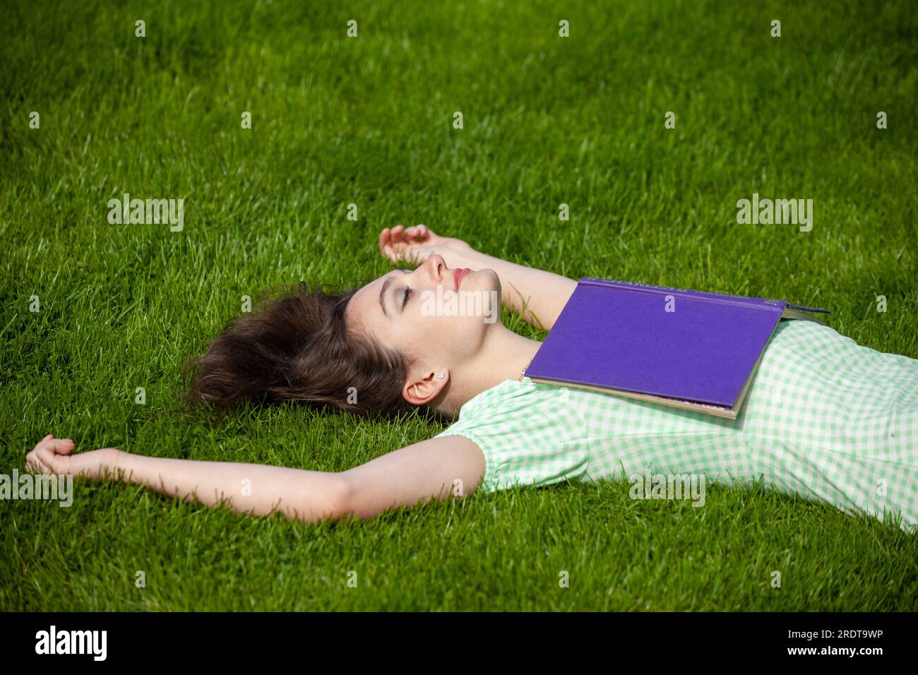A young woman lying on her back with her arms outstretched. She is wearing a summer dress and has a book on her chest. Stock Photo