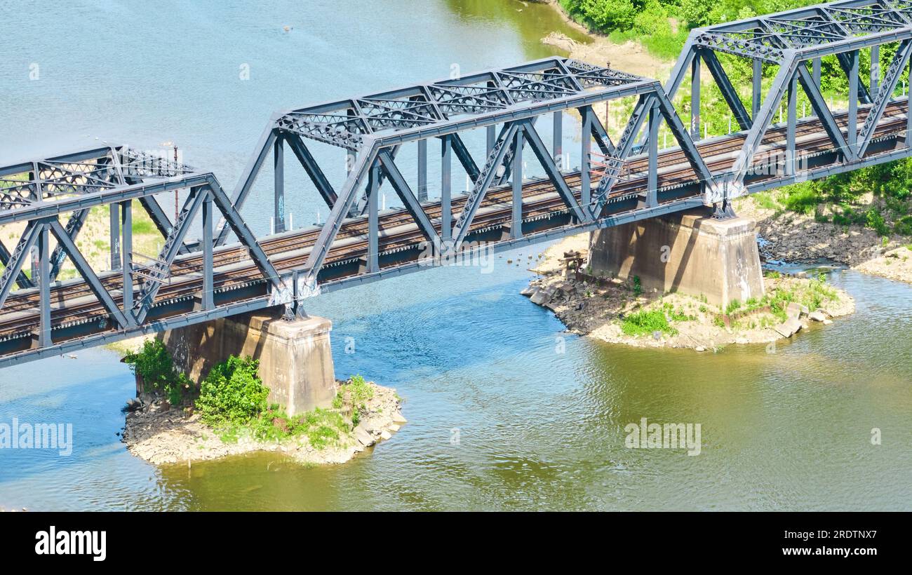 Aerial blue metal bridge with dual train tracks over river water leading to green bank aerial Stock Photo