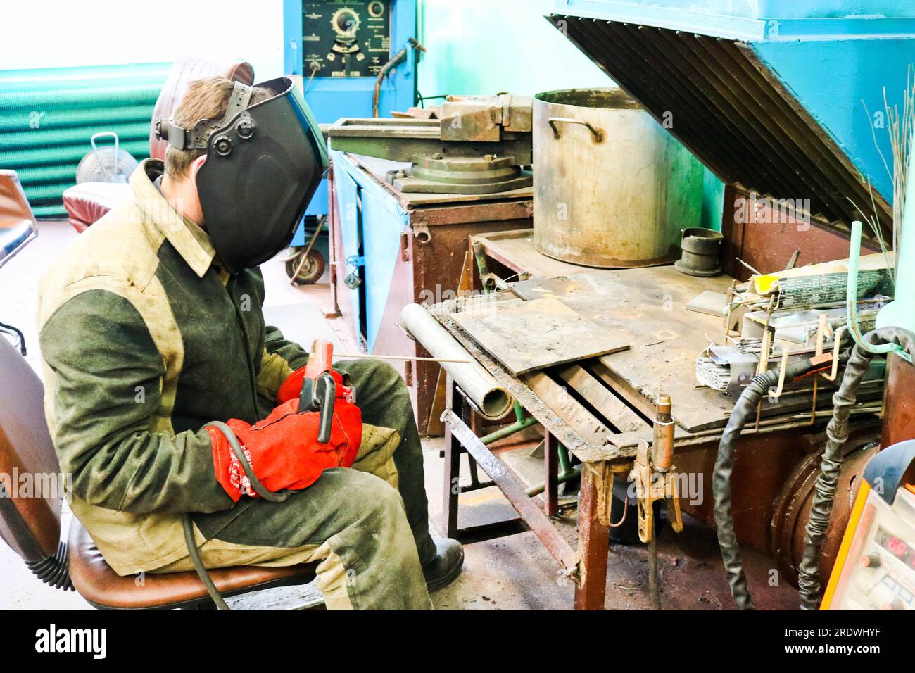 A male worker a welder in a protective mask welds a metal pipe at a welding station in a workshop at a metallurgical plant in a repair facility. Stock Photo