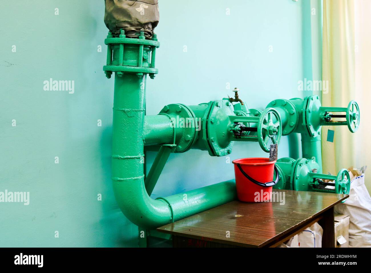 Leaking faulty valve, fitting on the pipeline with a bucket under it in the factory plant. Concept: repair of pipe and valve with available tools. Stock Photo