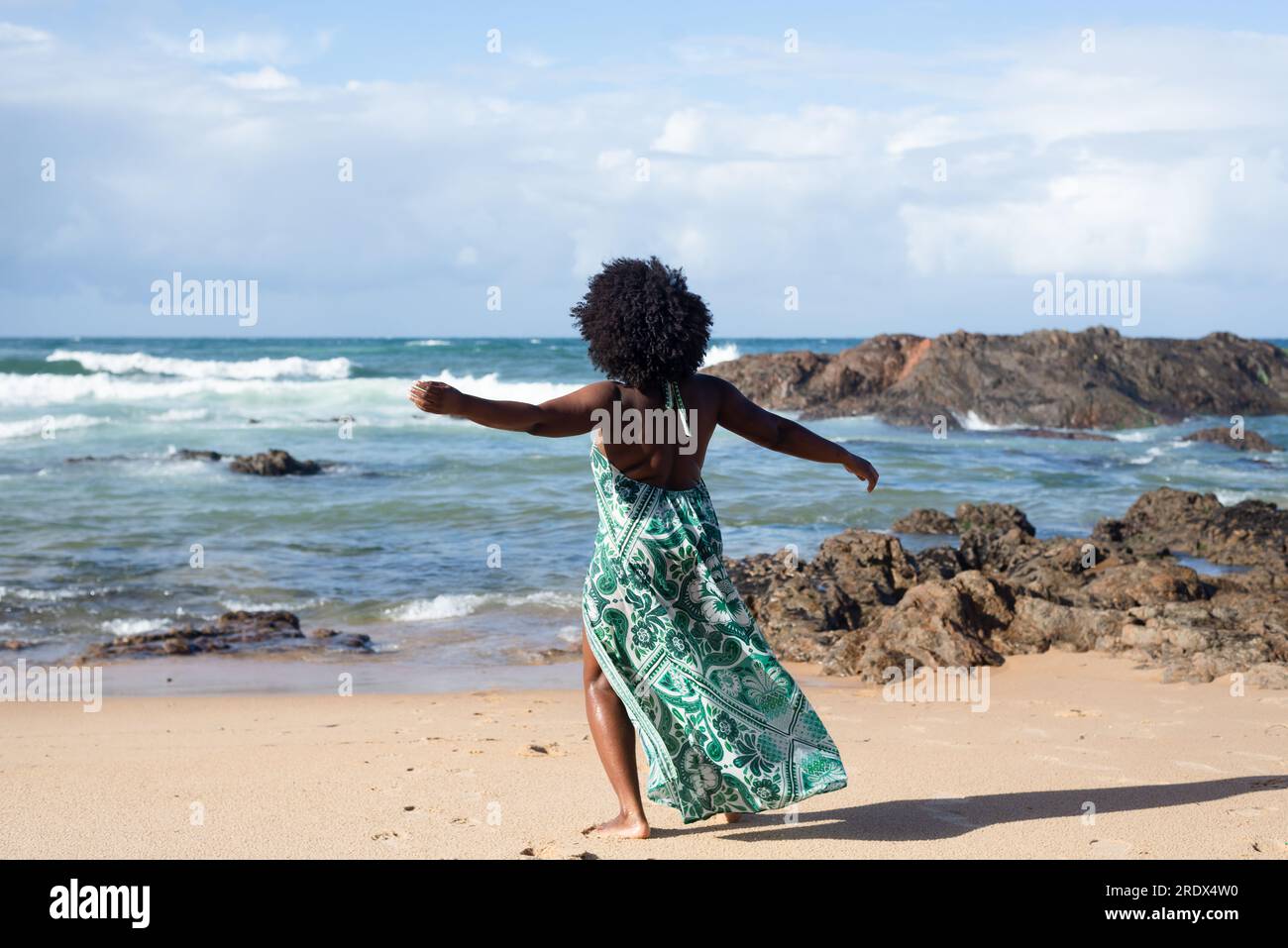 Full length portrait of a beautiful woman with black power hair standing on the beach sand with her back made movements with her arms. Sky clouds rock Stock Photo
