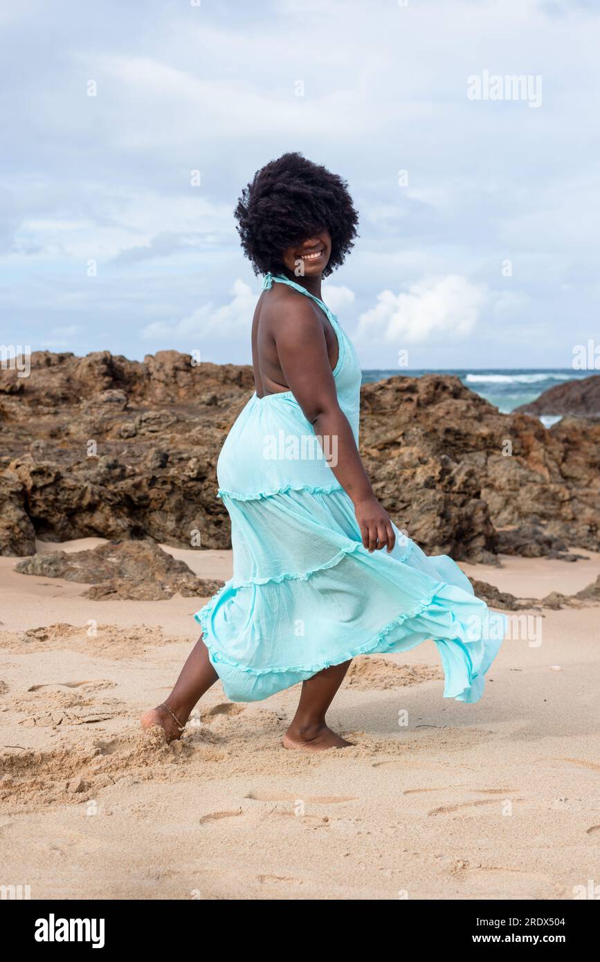 Beautiful woman with black power hair wearing long light blue outfit dancing on the beach sand. Happy and confident woman. Sky and clouds in the backg Stock Photo