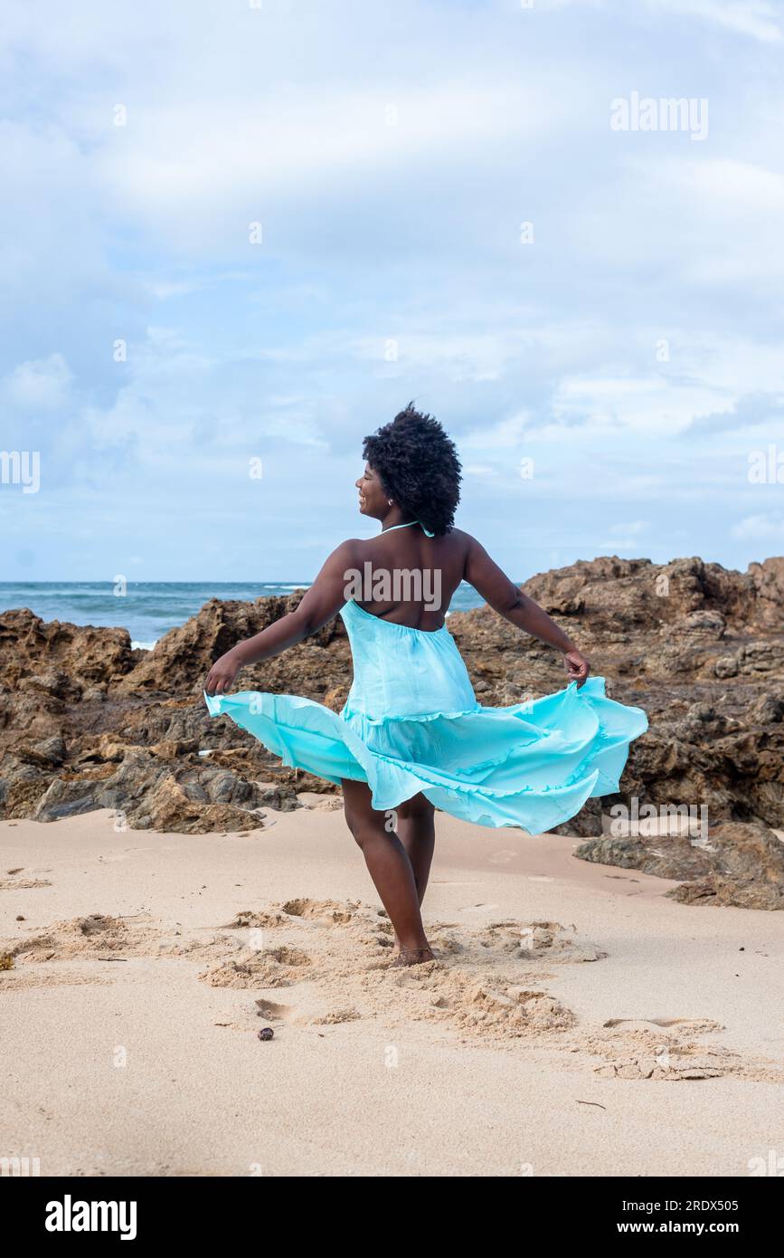 Beautiful woman with black power hair wearing long light blue outfit dancing on the beach sand. Happy and confident woman. Sky and clouds in the backg Stock Photo