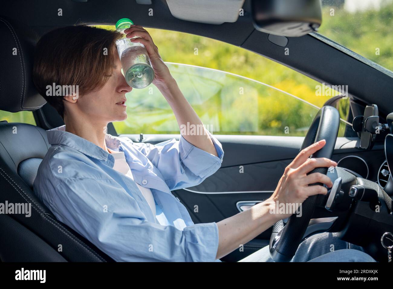 Hot weather for drivers. Tired from heat woman in car take break cooling forehead with cold bottle  Stock Photo