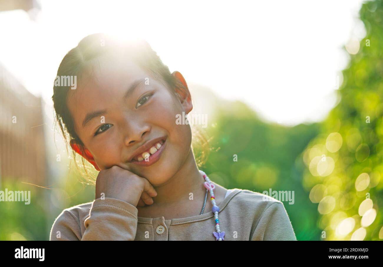 Portrait of healthy child girl, Asian child girl at 9-10 years old on vacation, sunlight flare behind, bokeh of green garden, eyes looking at camera, Stock Photo