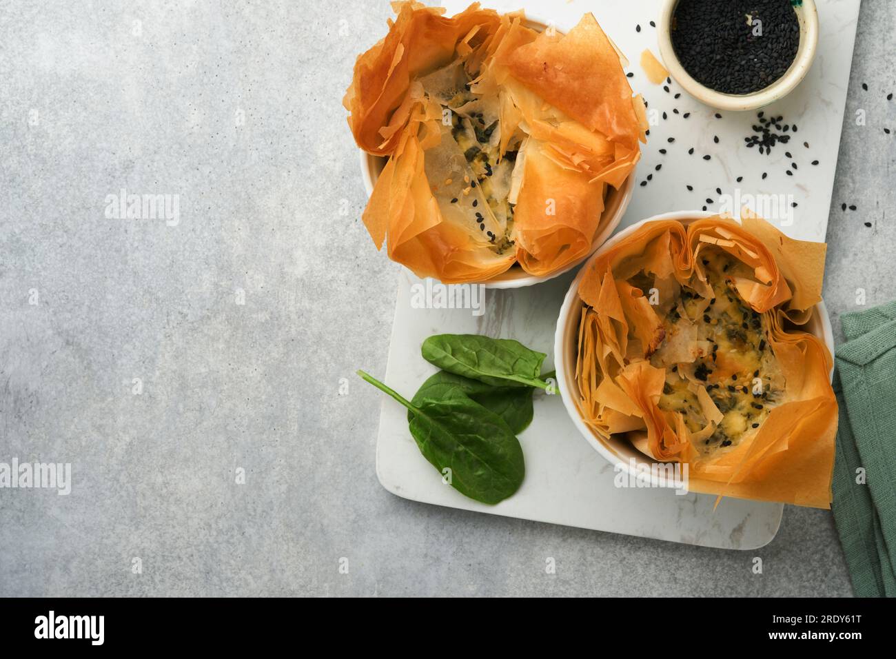 Filo pies with soft feta cheese and spinach in ceramic molds on old grey table background. Filo portions pies. Small Baked Spanakopita pies. Top view. Stock Photo