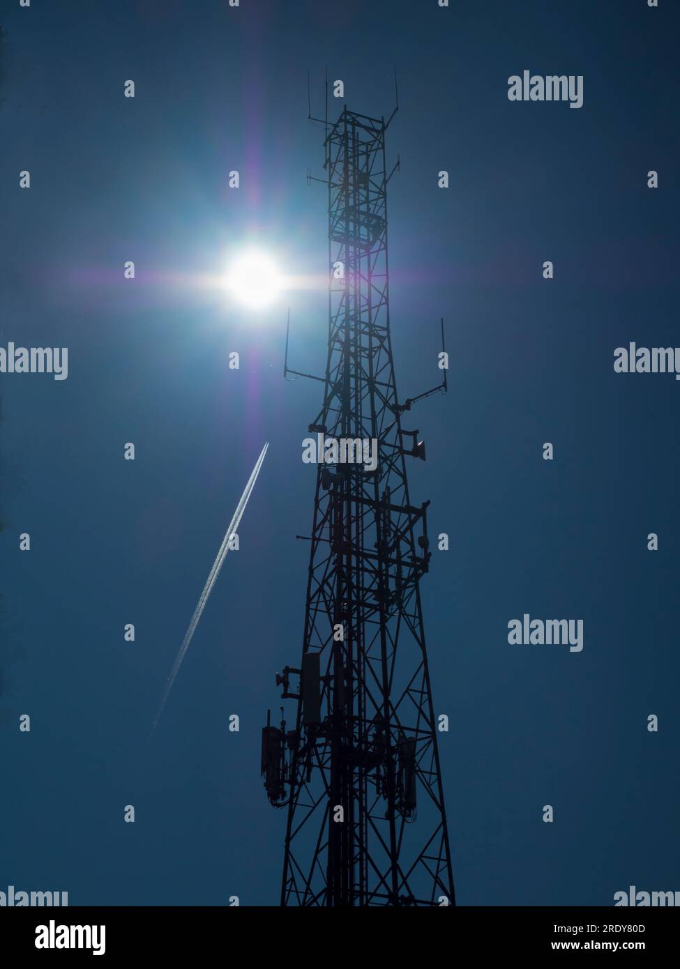 Headington, Oxford. An antenna-covered communications mast, silhouetted by the sun in a cloudless mid-summer sky, Far overhead, a passing aircraft and Stock Photo