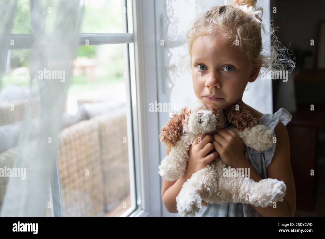 Sad girl holding stuffed toy standing near window at home Stock Photo