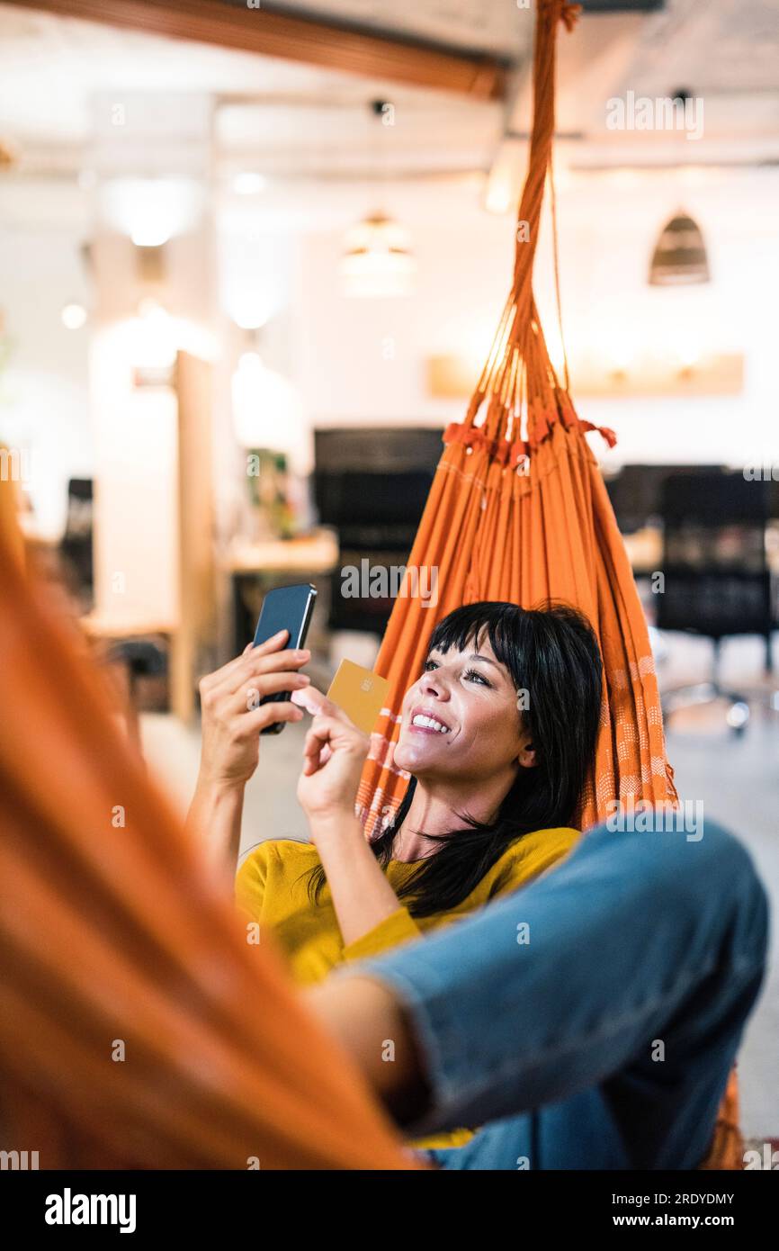 Happy mature businesswoman with credit card using smart phone in hammock Stock Photo