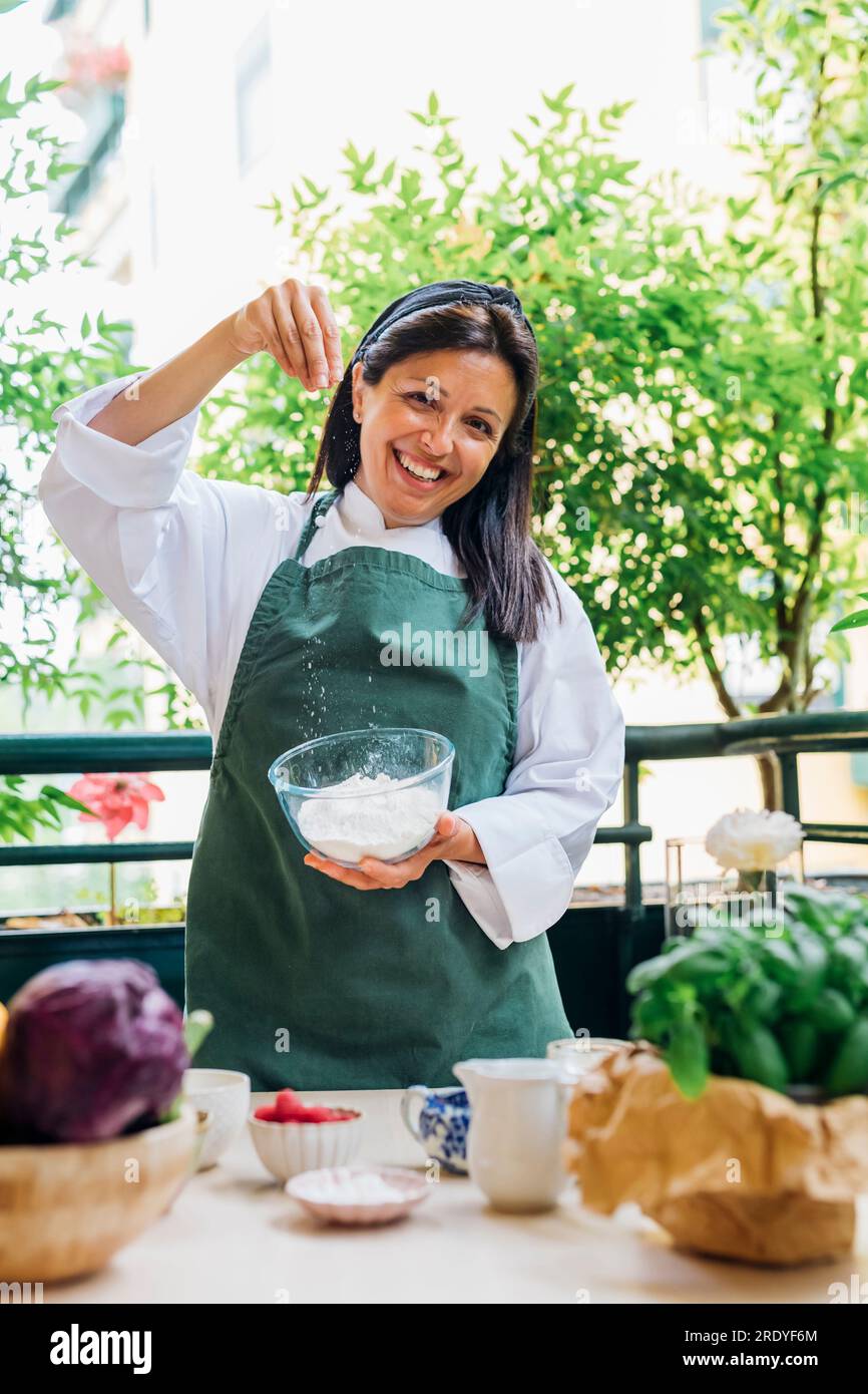 Happy woman holding bowl of flour standing by table in balcony Stock Photo