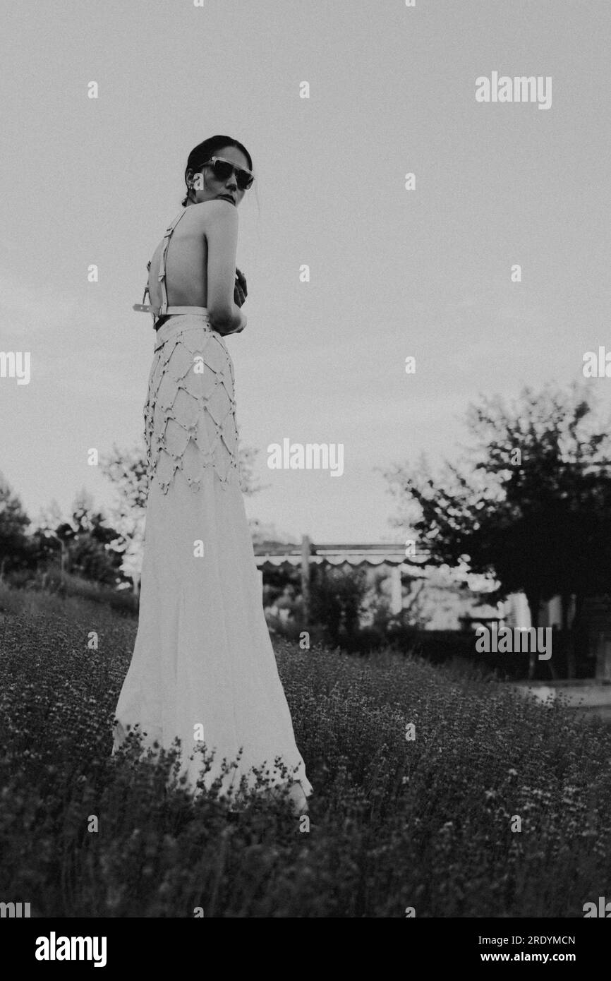 Beautiful young brunette woman in white dress and sunglasses posing in a lavender field at summer evening. Beauty concept. Stock Photo