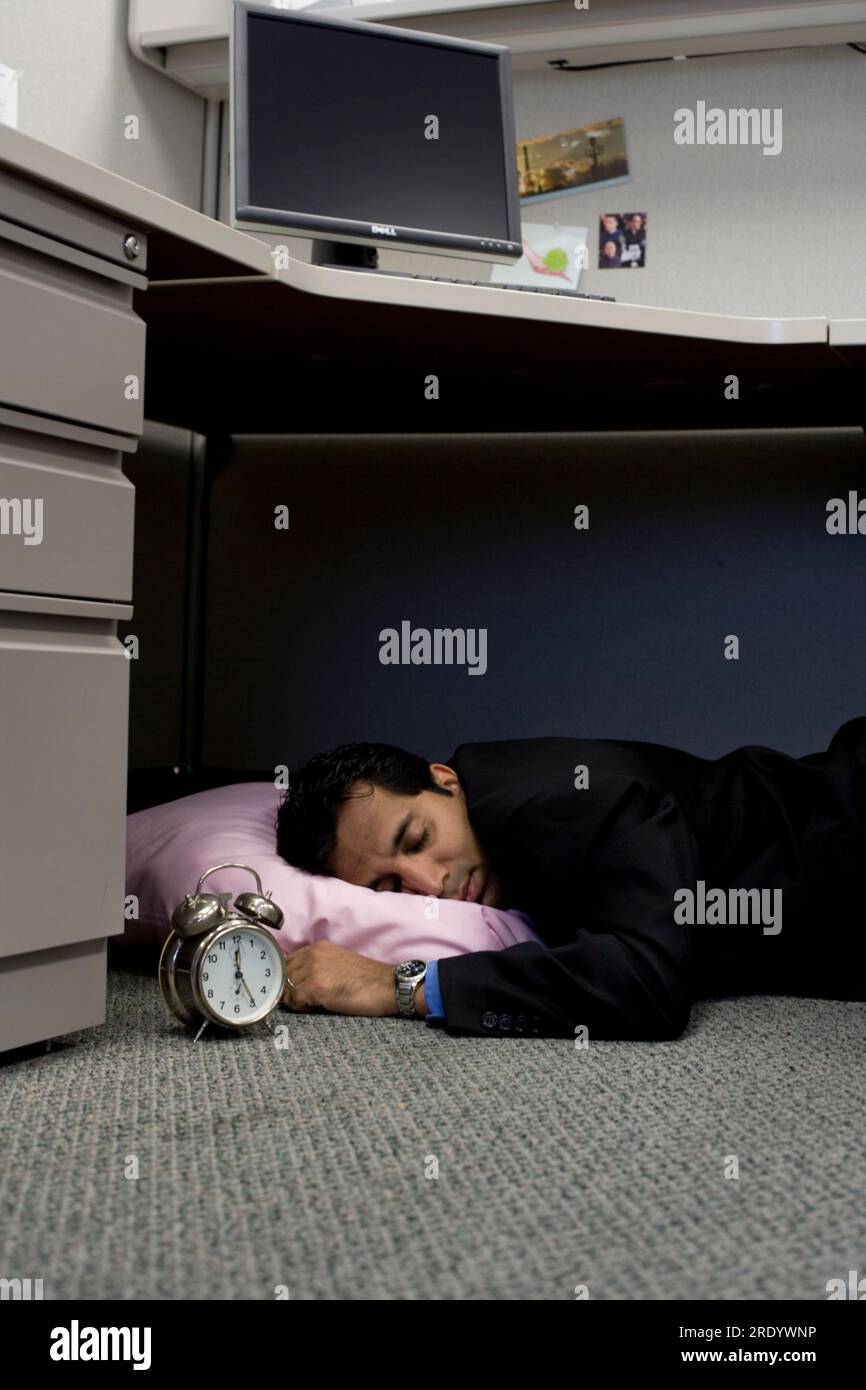 A man sleeping underneath his desk in his cubicle at work Stock Photo