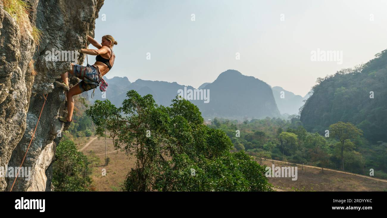 Female rock climber high on wall with trees and mountains behind, Asia Stock Photo
