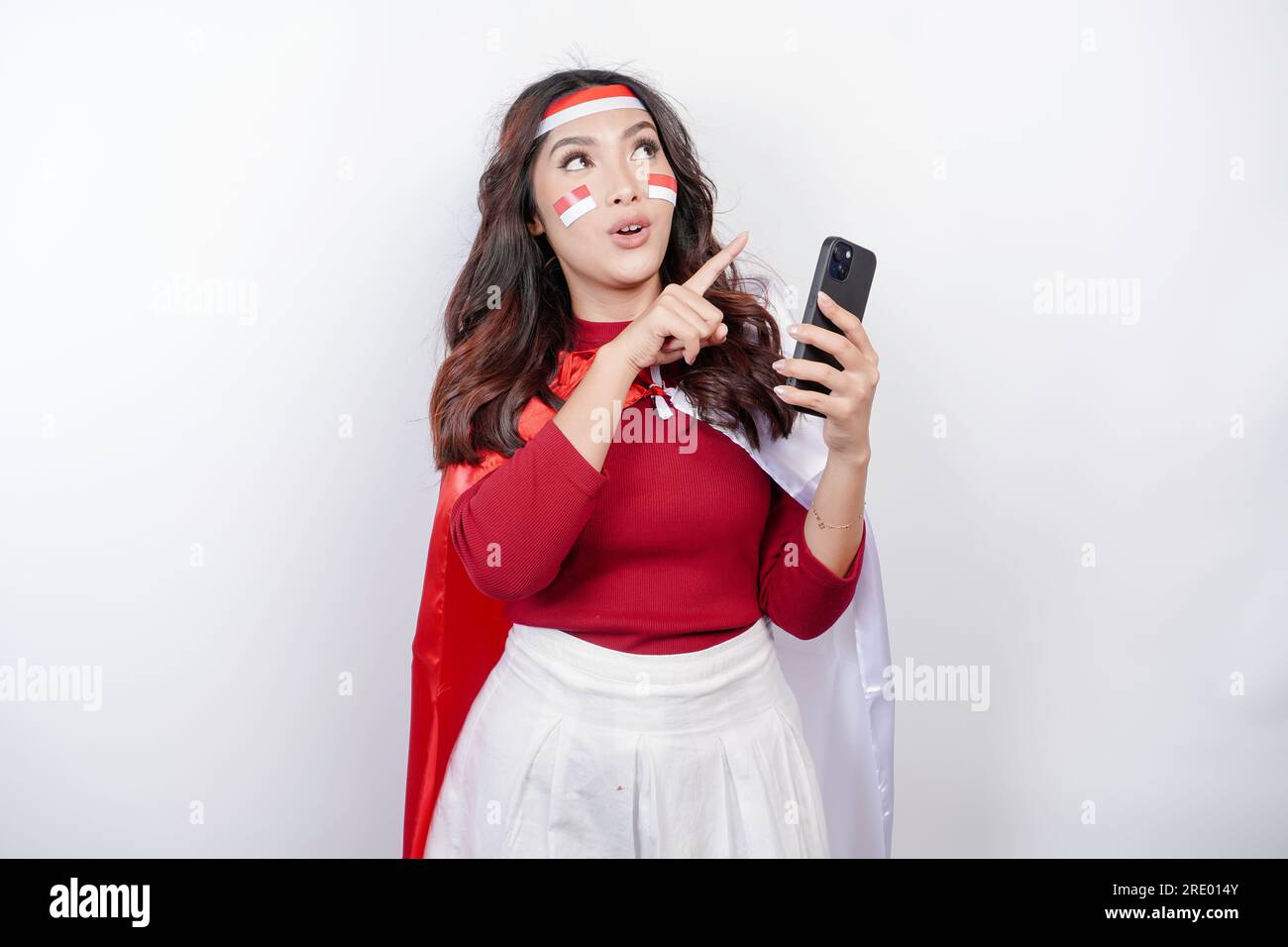 A portrait of a shocked Asian woman wearing headband, holding her phone, and pointing copy space on top of her, isolated by white background. Indonesi Stock Photo